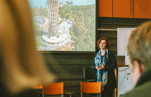 Hilde Sevens presenting to an audience, with an aerial render of a timber tower on screen behind her